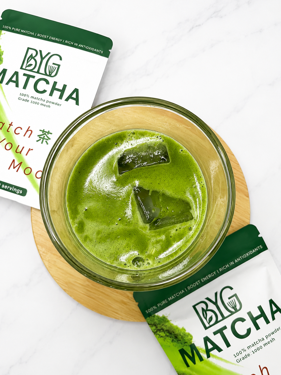 A glass bowl filled with bright green matcha tea sits on a round wooden coaster, with two BYG Matcha pouches placed behind and below it on a white marble surface.