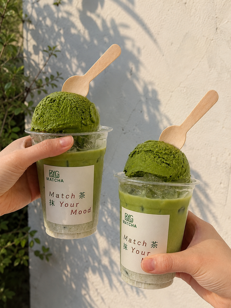 Two hands hold clear plastic cups of matcha green tea dessert outdoors against a white wall. Each cup contains a scoop of green matcha ice cream on top of a light green matcha drink, with a small wooden spoon and a white label that reads BIG MATCHA and Matcha Tea Your Mood.