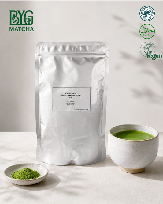 A silver stand-up pouch of BYG Matcha matcha powder stands on a light surface beside a small dish of green powder and a cup of green tea. The BYG Matcha logo appears in the top left, and certification with Rainforest Alliance, Halal  Certificate, Vegan. 