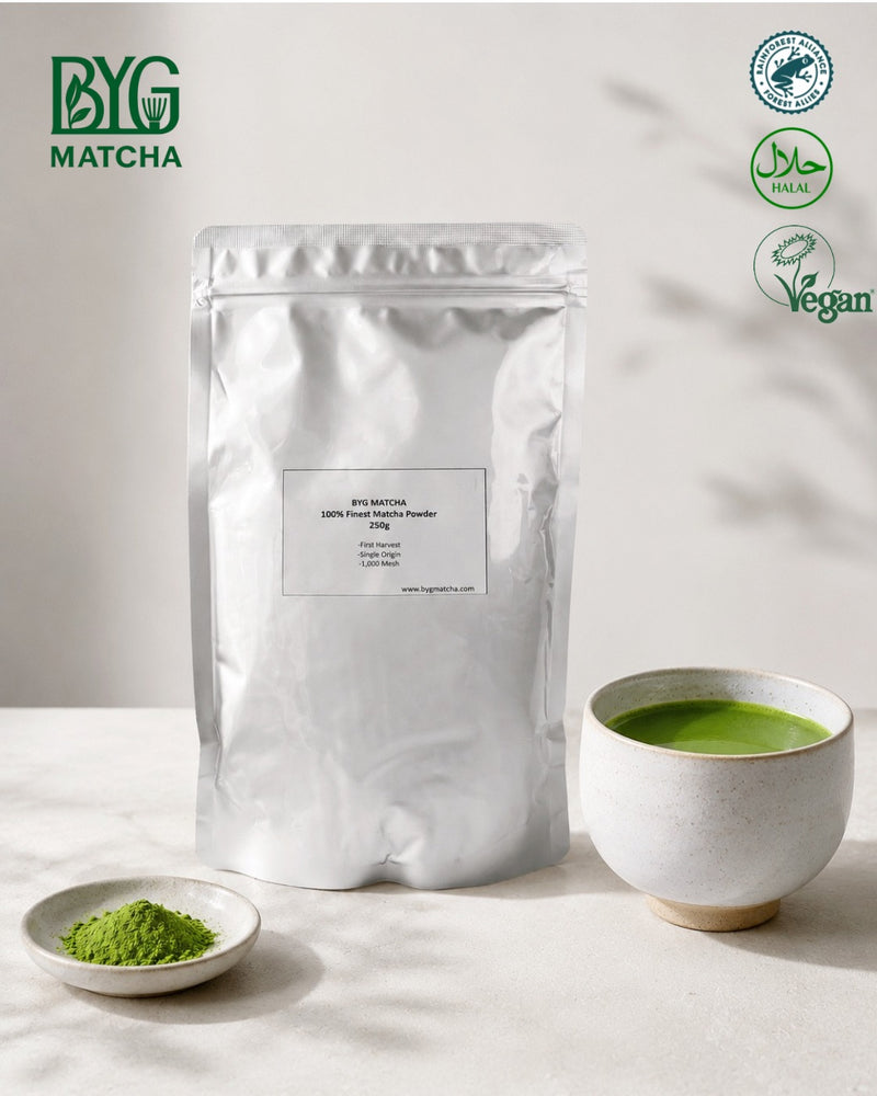 A silver stand-up pouch of BYG Matcha matcha powder stands on a light surface beside a small dish of green powder and a cup of green tea. The BYG Matcha logo appears in the top left, and certification with Rainforest Alliance, Halal  Certificate, Vegan. 