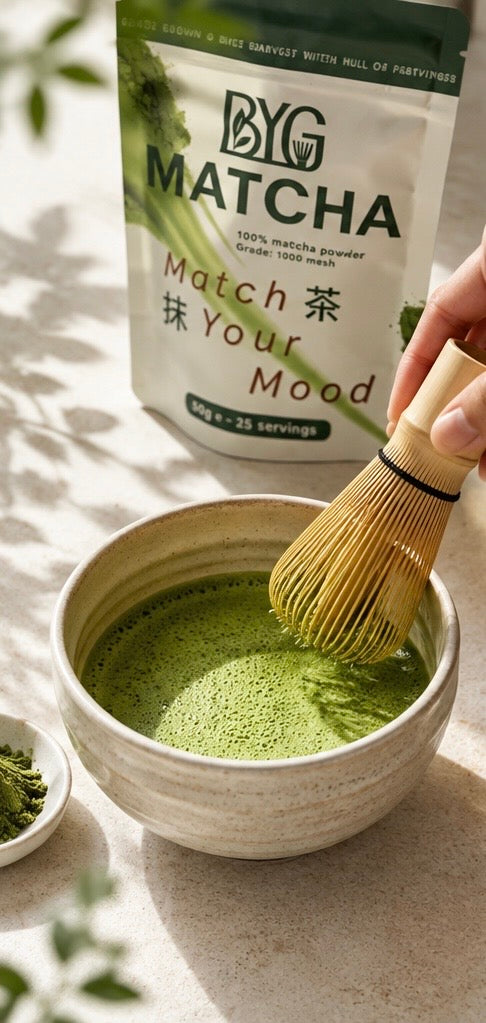 A hand holds a bamboo whisk over a bowl of green matcha tea, with a bag of BYG Matcha powder standing behind it on a sunlit surface.